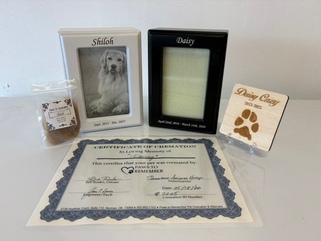 memorial package contents: engraved photo frame urns in white and black, a wooden paw print keepsake, a certificate of cremation, and a bagged clipping of hair